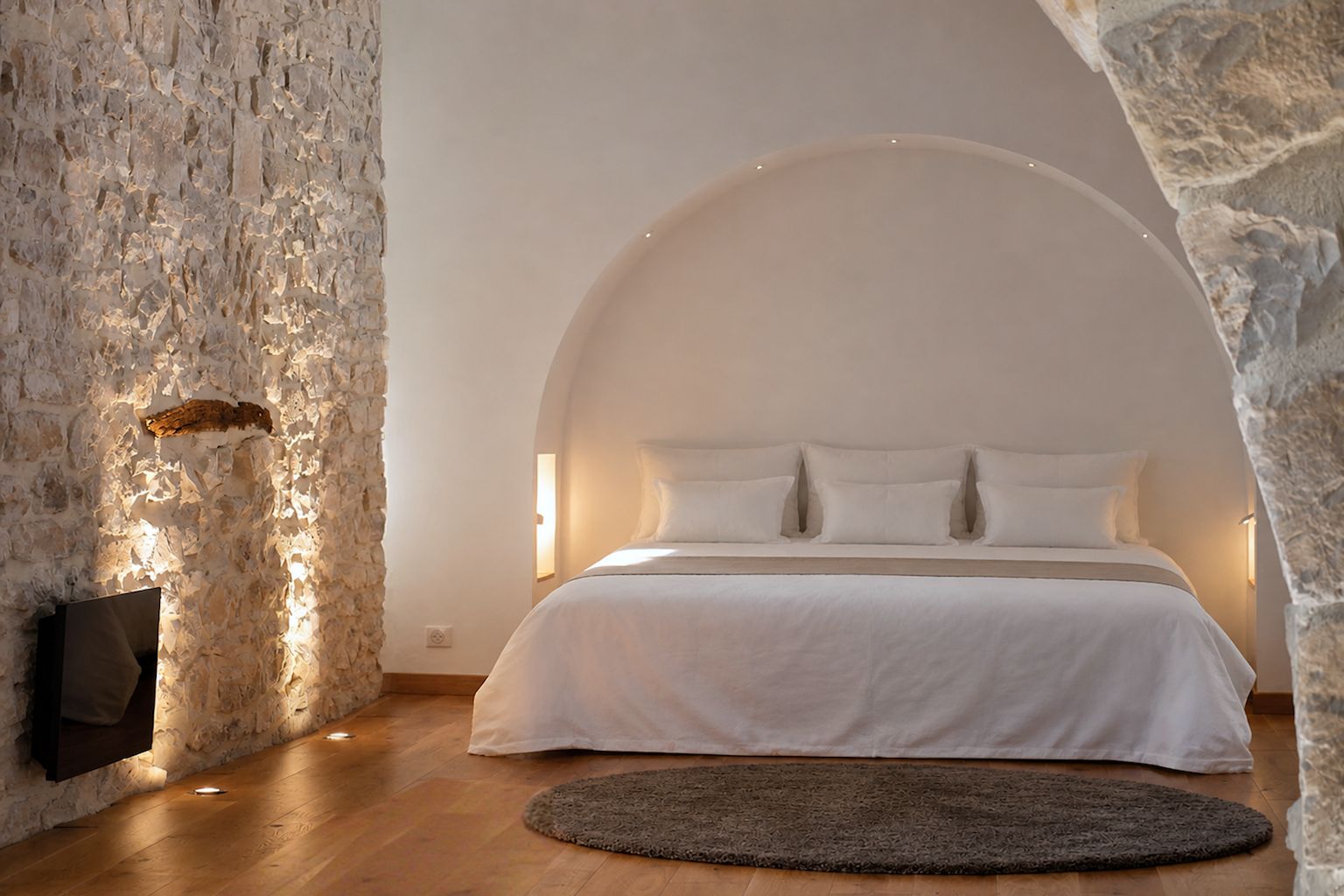 A bedroom with a stone wall, a bed with white bedding, and a round rug.