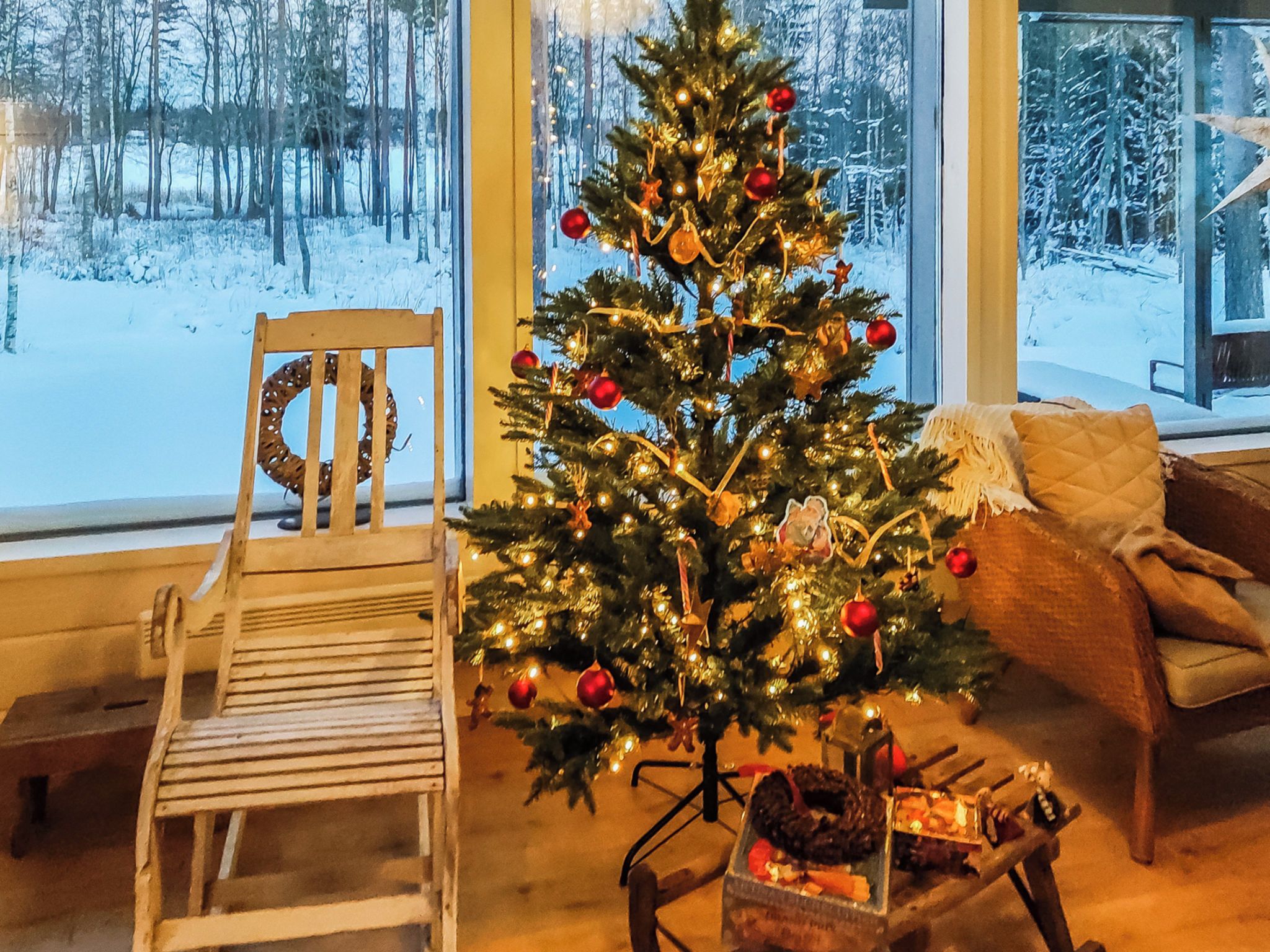 Una habitación acogedora con una silla de madera, un árbol de Navidad y vistas a un paisaje nevado a través de amplias ventanas.