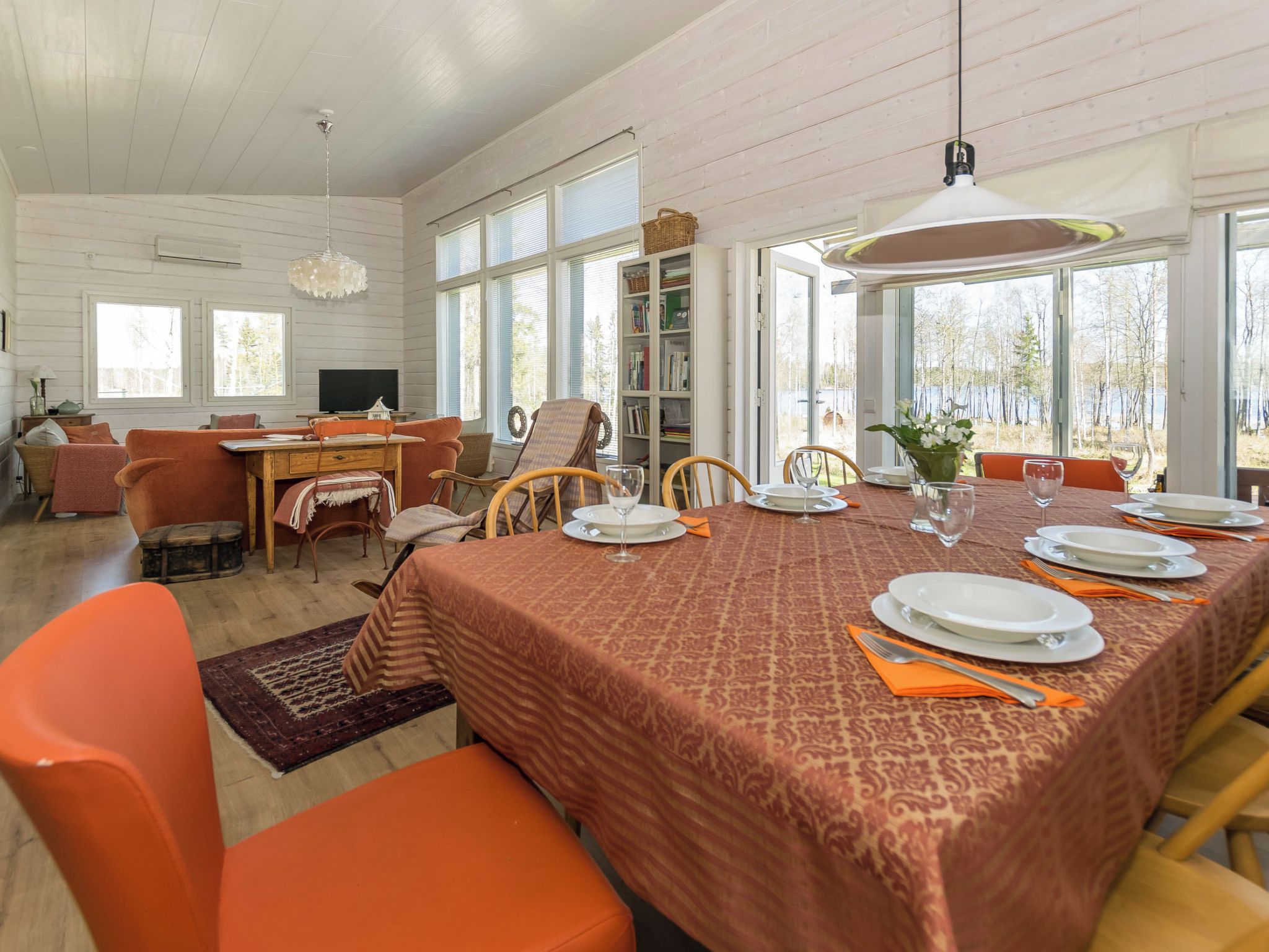 A dining area with a table set for four, surrounded by orange chairs, a bookshelf, and a television in the background.