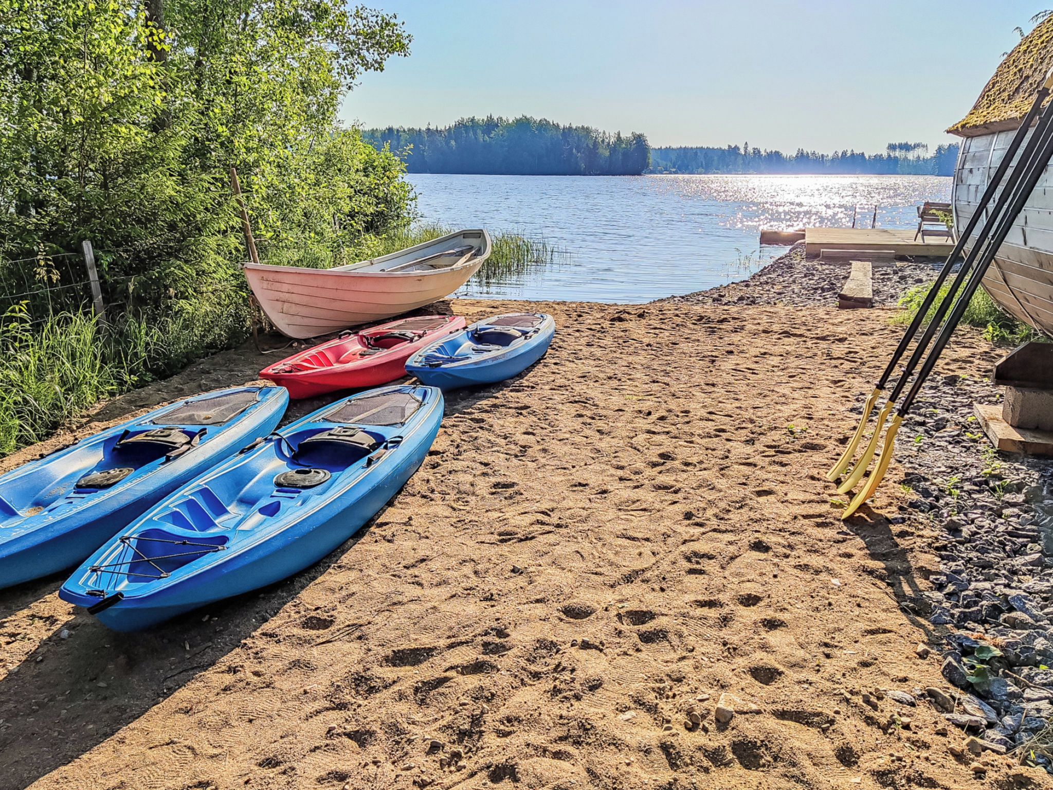 A sandy beach with kayaks and a boat, a wooden fence, and a forest in the background.