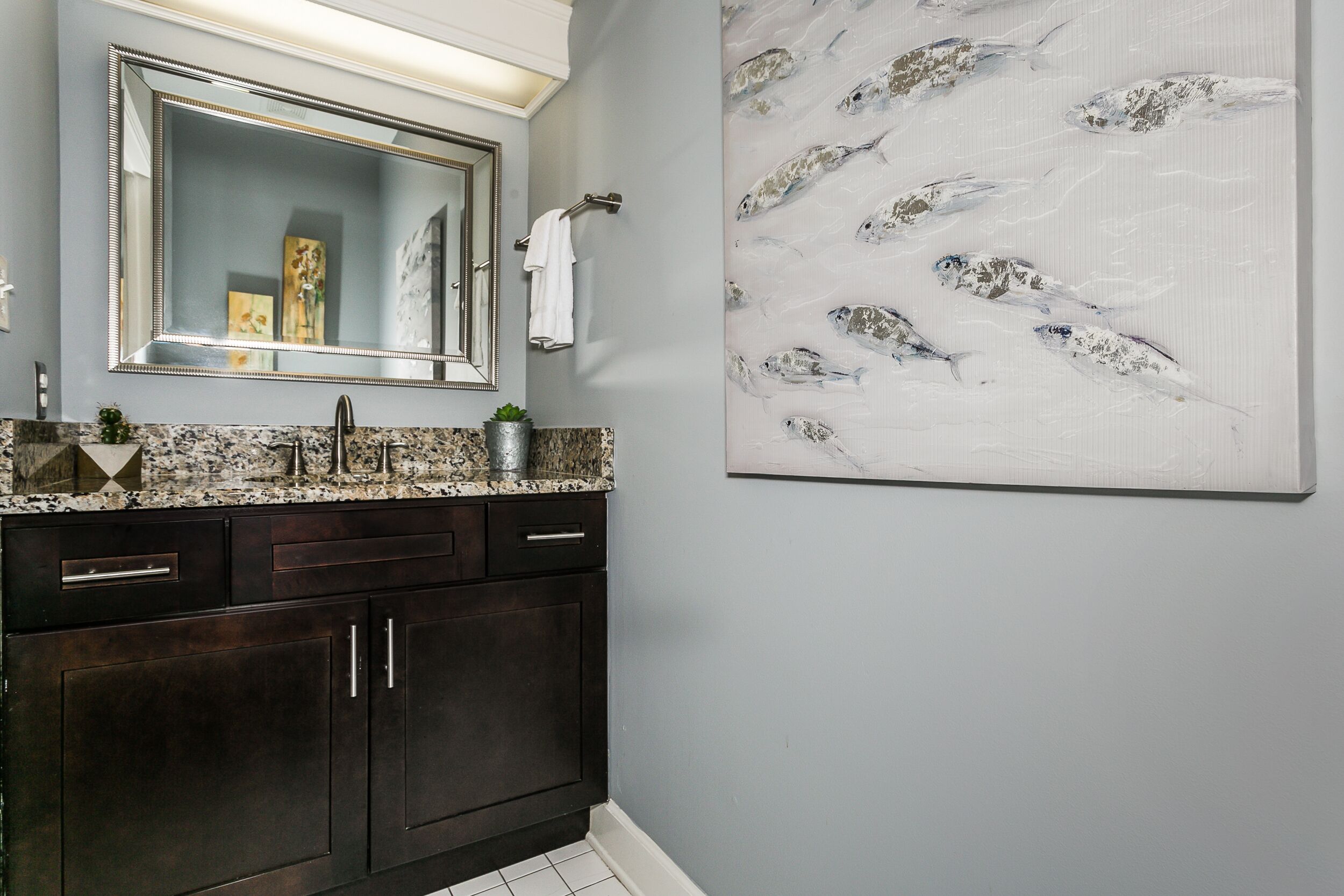 A bathroom with dark wooden cabinets, a granite countertop, a large mirror, and a wall-mounted artwork of fish.