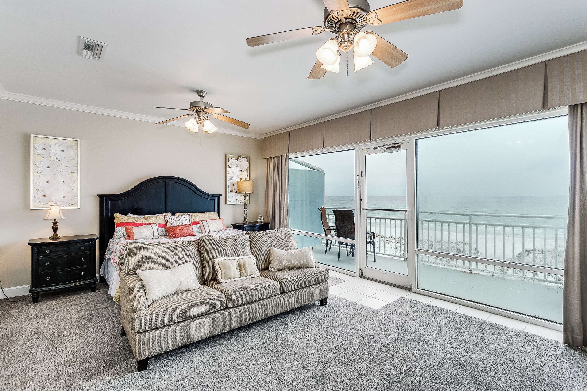 A bedroom with a large bed, a sofa, a nightstand, and a ceiling fan. There is a balcony with a view of the ocean.