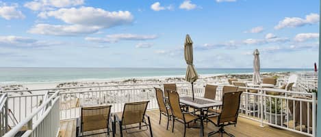 Una terraza de madera con asientos exteriores, barandillas blancas y vistas a la playa.