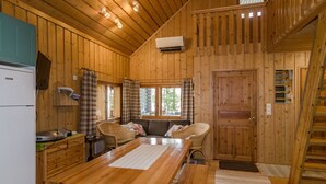 Interior de una cabaña de madera con una mesa de comedor, sillas, un sofá, una zona de cocina con refrigerador y una ventana con cortinas.