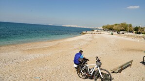 Une plage avec un vélo garé dans le sable, une personne assise sur un banc et une vue dégagée sur la mer et les montagnes lointaines.
