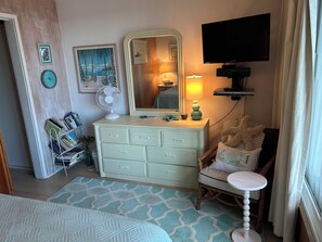 Ein Schlafzimmer mit Kommode, Stuhl, Bett, Fernseher und einem Fenster mit Vorhängen.