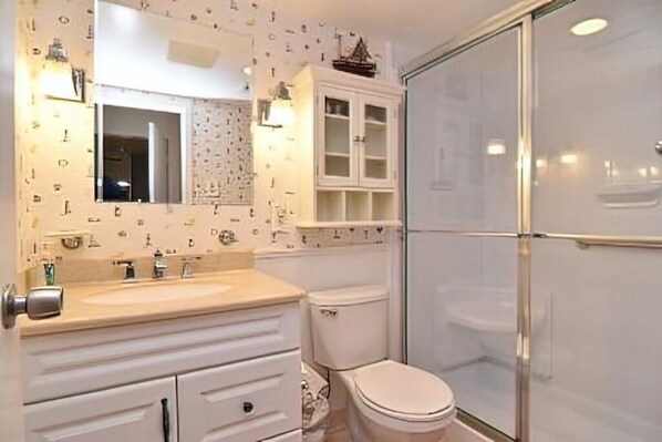 Un baño con lavamanos blanco, espejo y mueble, inodoro y cabina de ducha con paredes de vidrio.