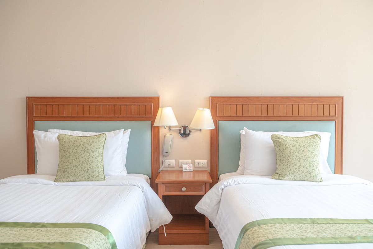 Une chambre d’hôtel avec deux lits, chacun doté d’une tête de lit en bois, de draps blancs et d’oreillers à motifs floraux verts. Une table de chevet se trouve entre les lits, avec une lampe et un téléphone.