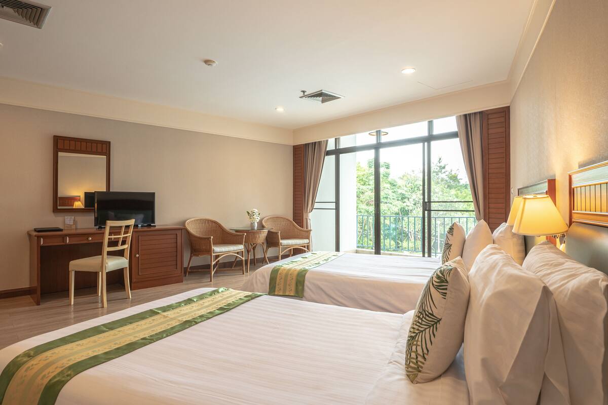 Une chambre d’hôtel avec deux lits, un bureau avec une télévision, une chaise et une vue sur la verdure grâce à de grandes fenêtres.