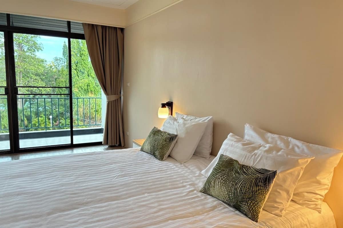 Une chambre d’hôtel avec un grand lit, deux oreillers, une table de chevet, une lampe et une vue sur la verdure grâce à de grandes fenêtres.