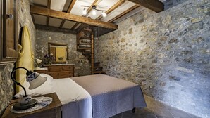 A bedroom with a stone wall, wooden beams, a bed, a dresser, a mirror, and a lamp.
