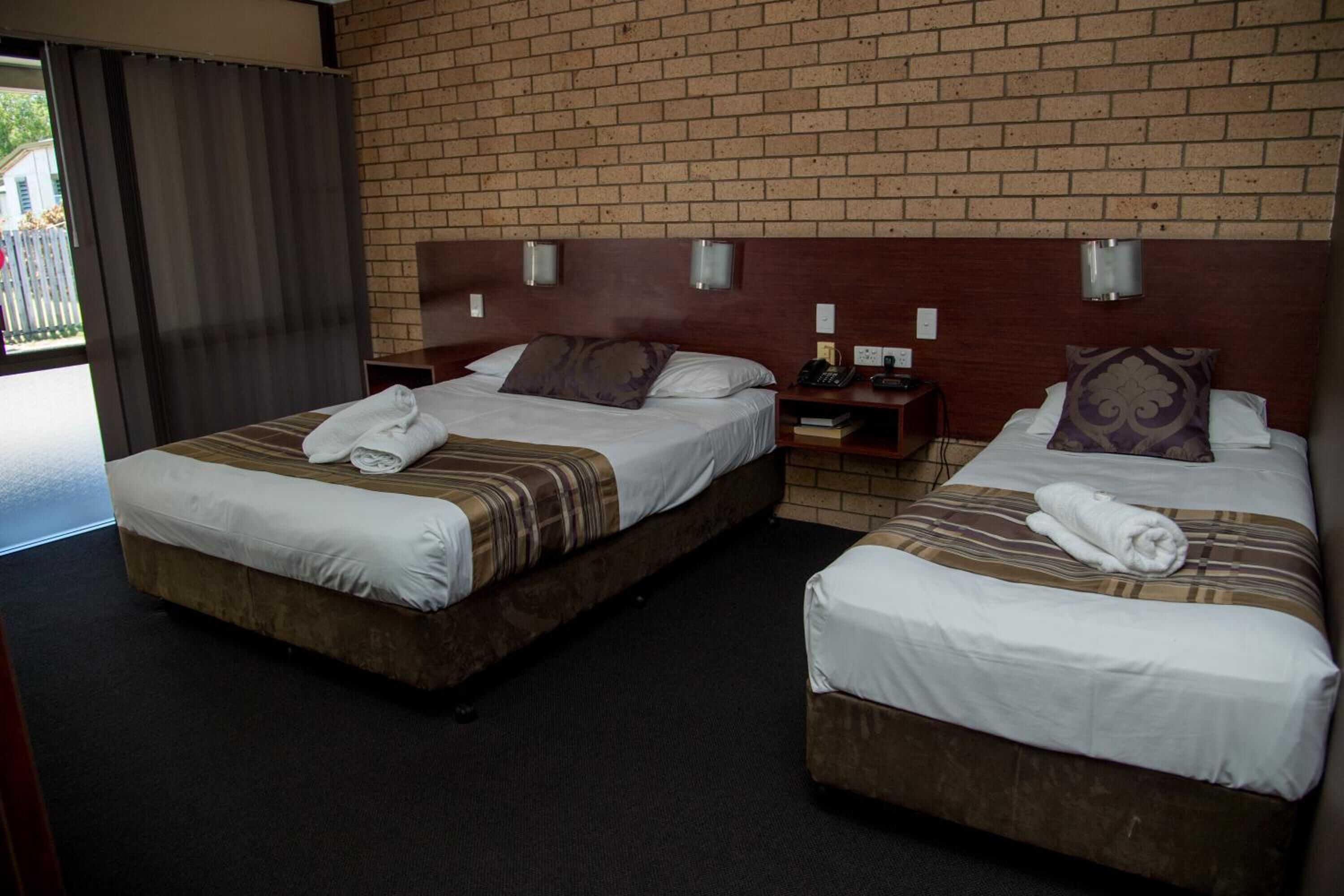 A hotel room with two beds, a brick wall, and a small table with a phone and a lamp.