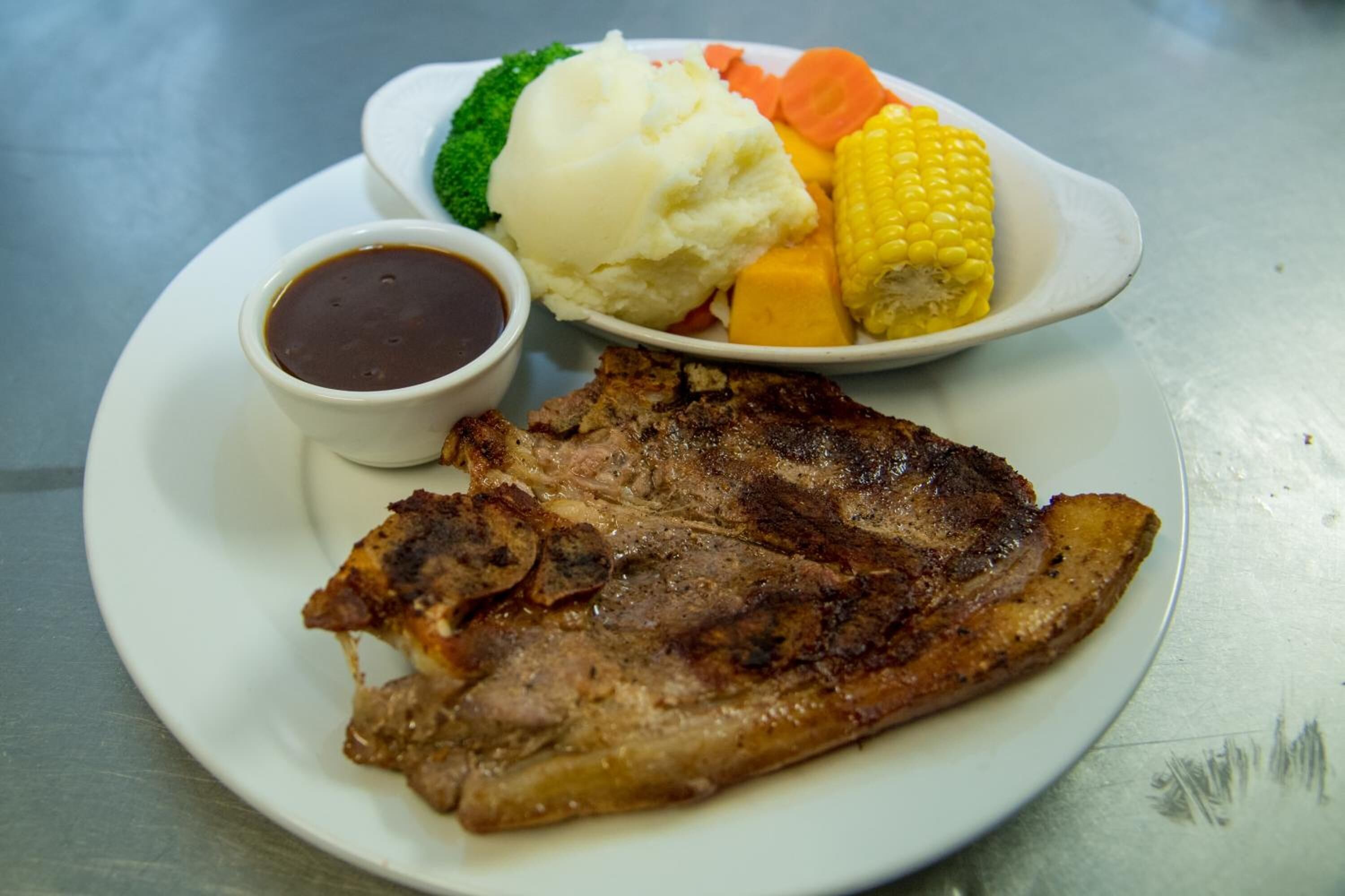 A plate with a grilled steak, mashed potatoes, corn on the cob, carrots, and a small bowl of sauce.