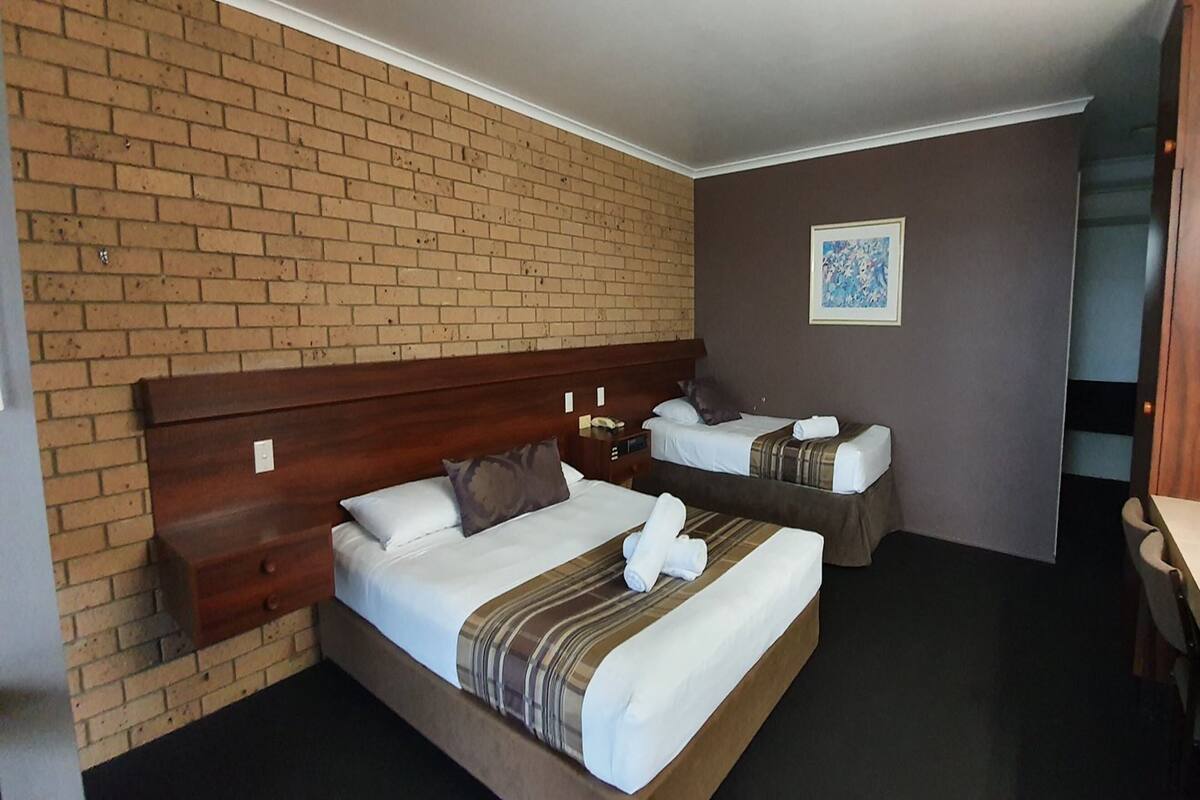 A hotel room with two beds, a wooden headboard, a small bedside table with a lamp, and a painting on the wall.