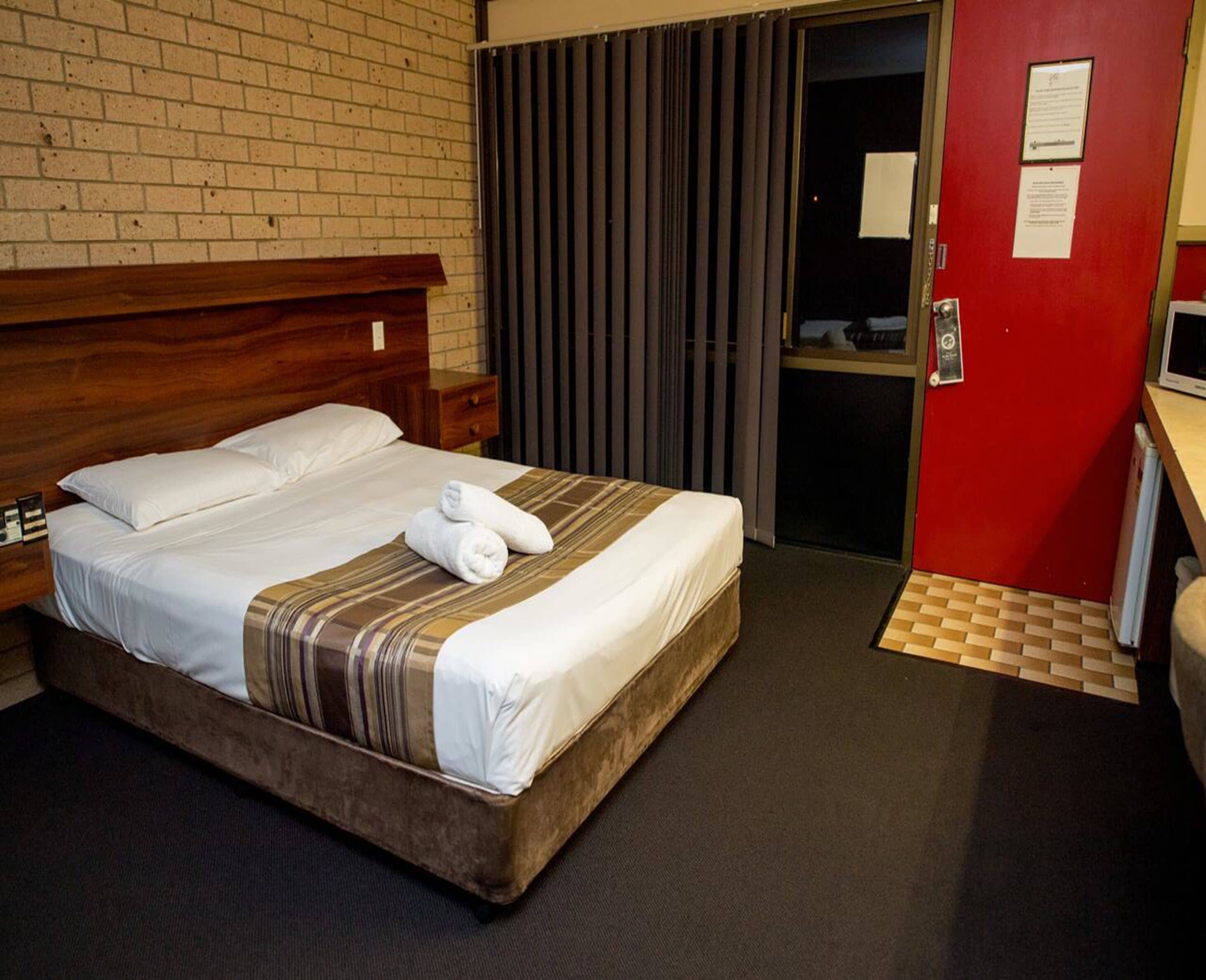A hotel room with a wooden headboard, a bed with white linens and a striped blanket, a red wall, and a door with a mirror.
