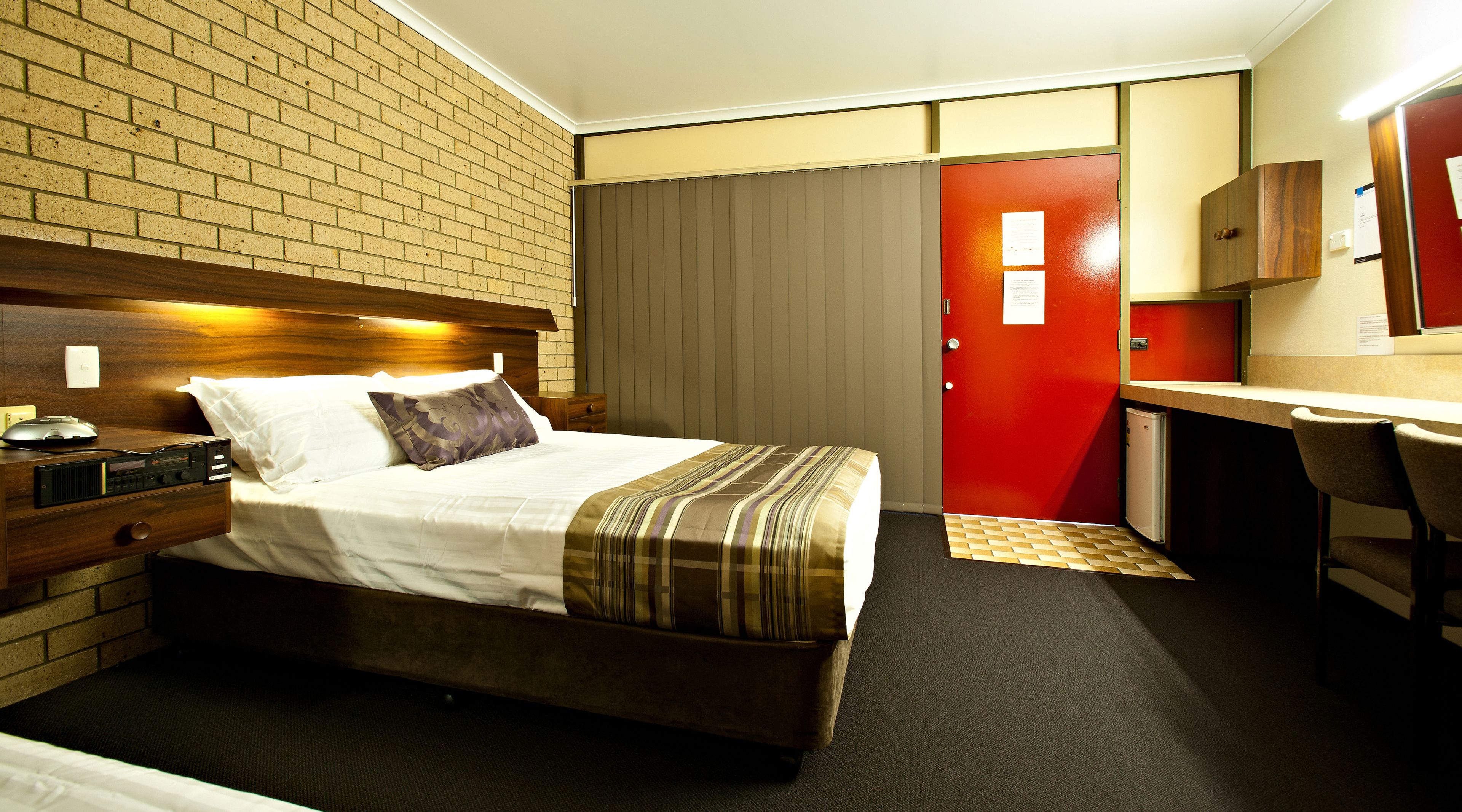 A hotel room with a bed, bedside table, desk, chair, and a red door.