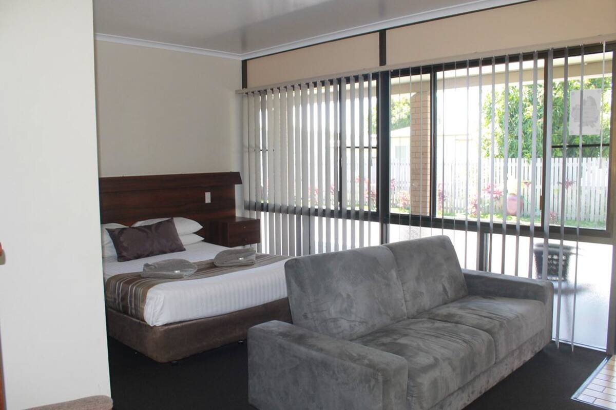 A bedroom with a bed, a grey sofa, and a window with blinds.