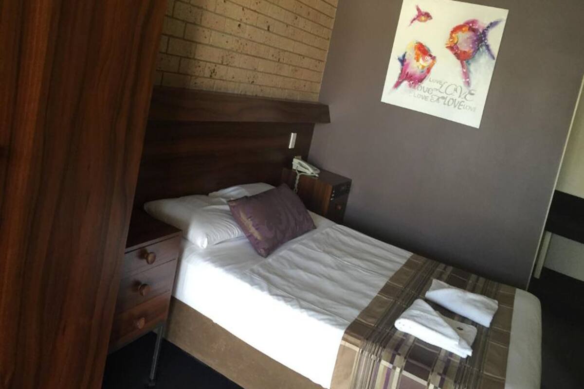 A neatly made bed with a white comforter and a plaid blanket, a wooden headboard, a nightstand with a phone, and a wall with a colorful fish painting.