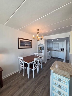 A dining area with a wooden table and chairs, a chandelier, and a kitchen in the background.