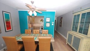 A dining area with a table set for four, a ceiling fan, and a kitchen in the background. - WALK RIGHT ONTO THE BOARDWALK FROM APARTMENT PATIO! (Rehoboth Beach)