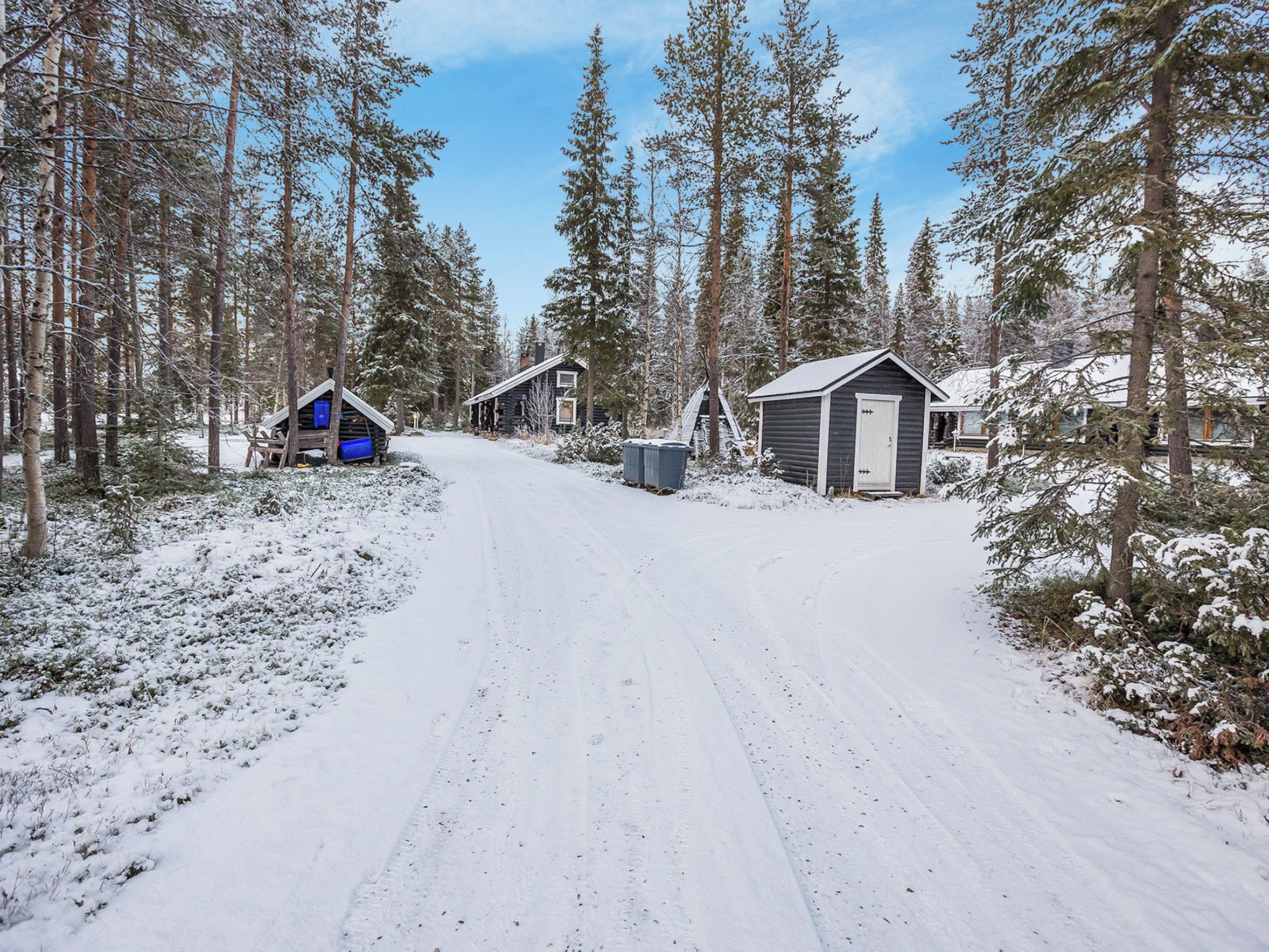 Un camino cubierto de nieve que conduce a una zona con cabañas, con varios edificios, incluyendo un cobertizo pequeño y una cabaña más grande con una puerta.