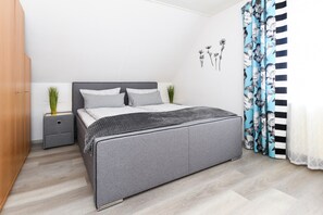Quarto com cama cinza, mesa de cabeceira, guarda-roupa e janela com cortinas.