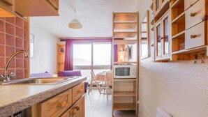 A compact kitchen with wooden cabinets, a built-in microwave, and a window with curtains. - Soyouz Vanguard by Interhome (Le Corbier)