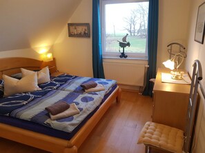 Ein ordentlich bezogenes Bett mit einer gemusterten Decke, einer Nachttischlampe und Blick auf einen Garten durch ein Fenster.