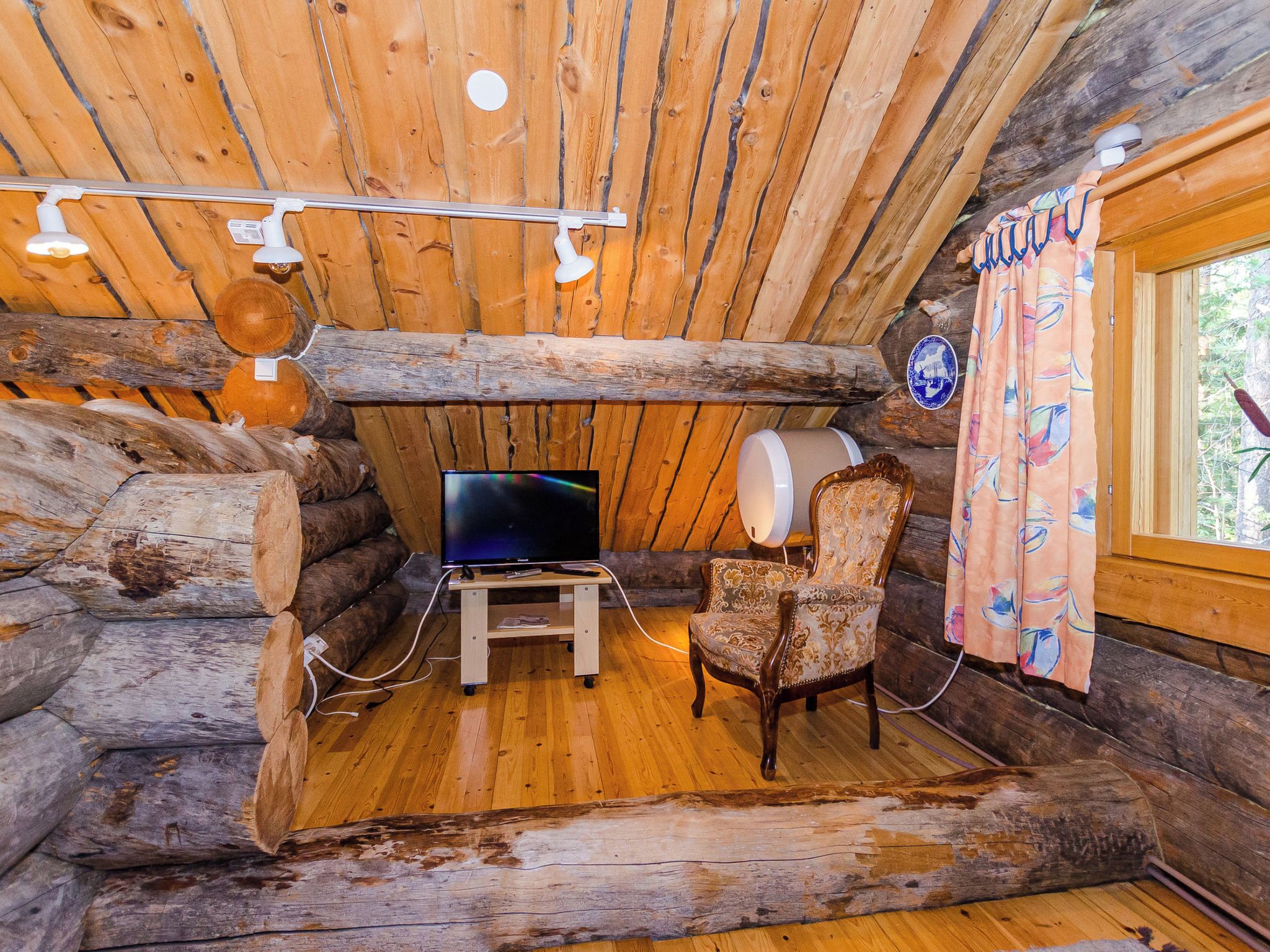 Interior de cabana de tronco com televisão em móvel, uma cadeira e janela com cortinas.