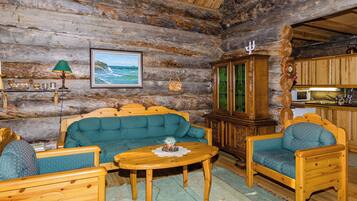 A log cabin interior with a wooden sofa, armchairs, a dining table, and a kitchen area.