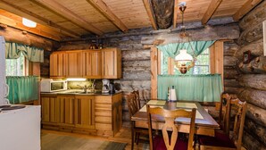 L’intérieur d’un chalet rustique est doté d’une table à manger en bois, de chaises, d’un coin cuisine avec des placards en bois, d’un réfrigérateur blanc et d’un luminaire suspendu.