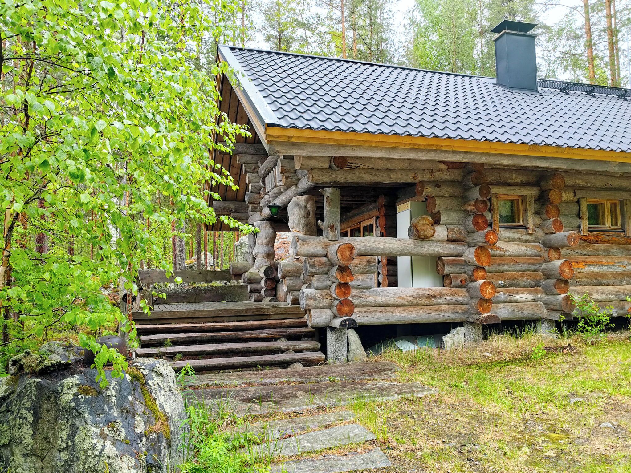 En timmerstuga med ett tak täckt av tegelpannor, en trätrappa som leder upp till entrén och en skog som bildar bakgrunden.
