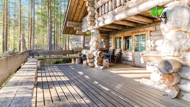 Une cabane en rondins dotée d’une terrasse en bois, d’un coin repas avec des sièges extérieurs et d’une grande fenêtre donnant sur la forêt.