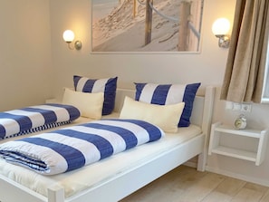 Una cama doble con ropa de cama a rayas azules y blancas, dos lámparas de pared, un cuadro de un paisaje nevado, una mesita de noche con un reloj y una ventana con cortinas.
