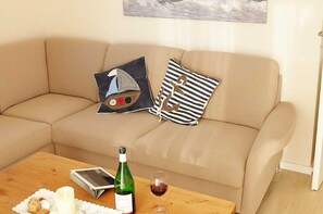 Un divano color beige con due cuscini decorativi, un tavolino in legno con una bottiglia di vino, un bicchiere di vino, una candela e un libro.