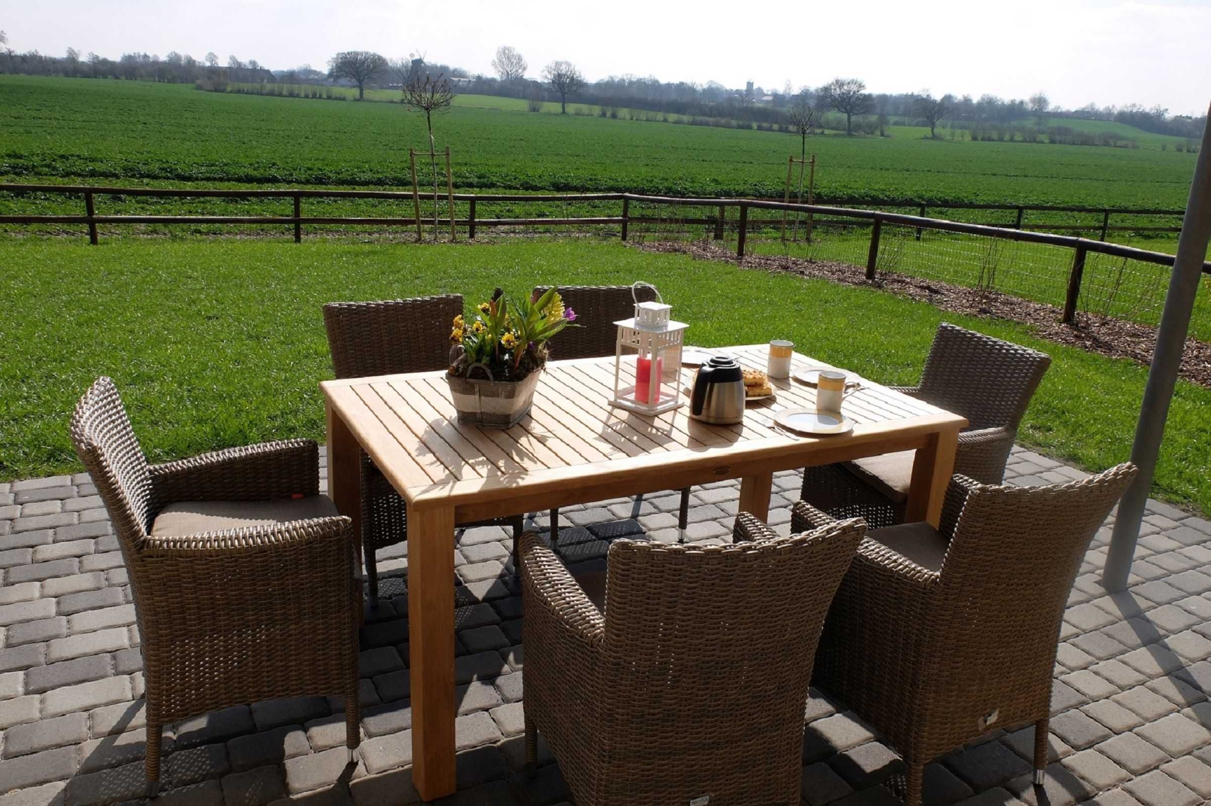 A wooden table with wicker chairs on a paved patio with a view of a green field.