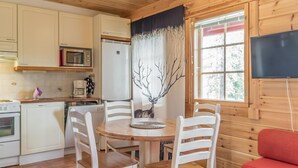 Una cocina acogedora de estilo rústico con armarios blancos, un microondas, un refrigerador y una mesa de comedor con cuatro sillas. Se aprecia un cuadro de un árbol en la pared y un televisor montado.