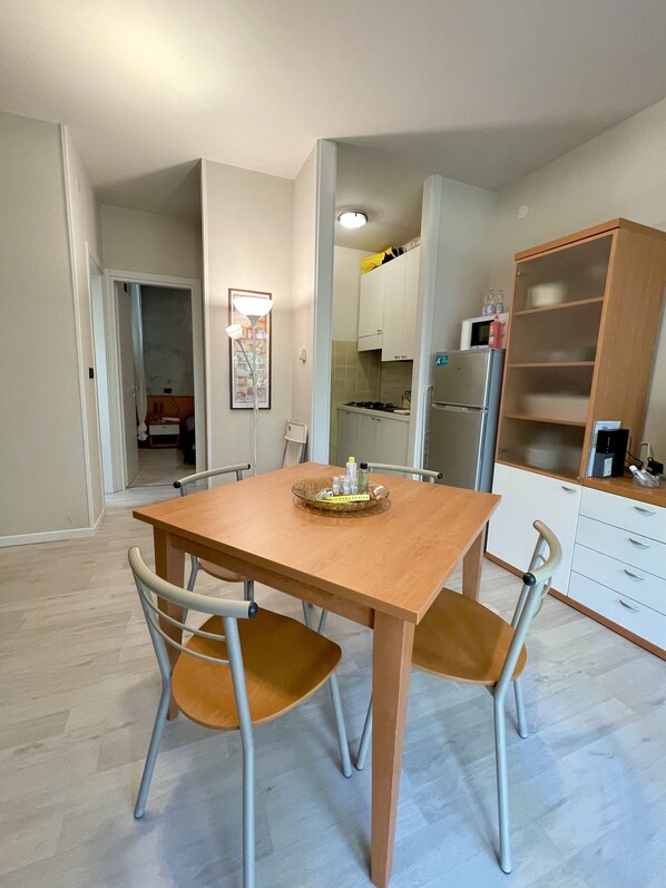A dining area with a wooden table and two chairs, a kitchen in the background, and a wooden cabinet with shelves.