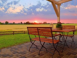 A patio with a brick floor, two wooden benches, a table with a potted plant, and a view of a field at sunset. - Cottage Country House - Ferienhof Börsby - Cottage Country house (Pommerby)