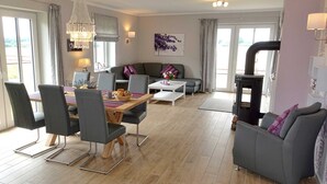 A modern living room with a dining area, grey sofas, a wooden dining table, and a fireplace. - Cottage Country House - Ferienhof Börsby - Cottage Country house (Pommerby)