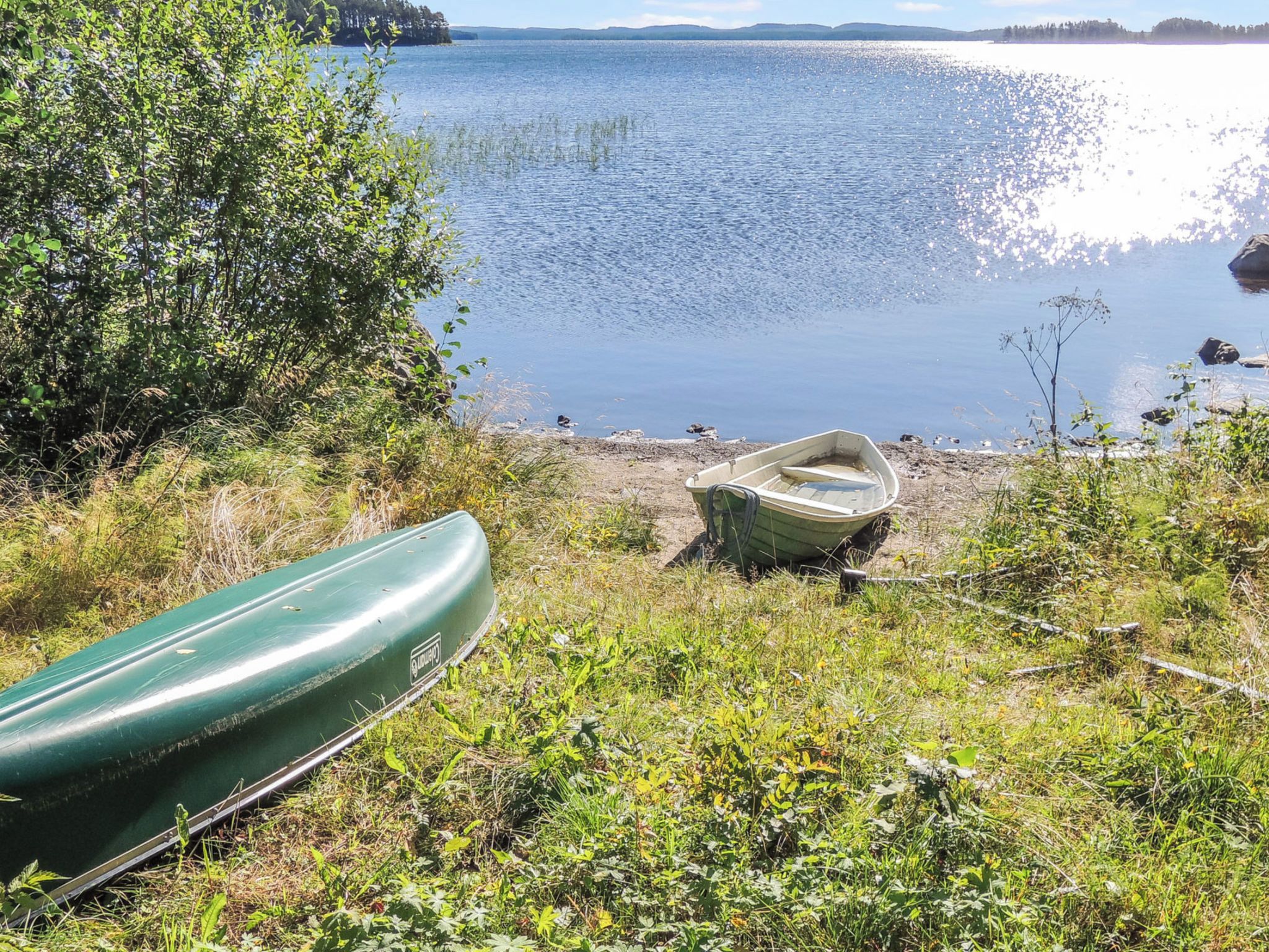Maisema järven rannalta, jossa näkyy rannalla oleva vene ja toinen vene, joka lepää ruohikolla.