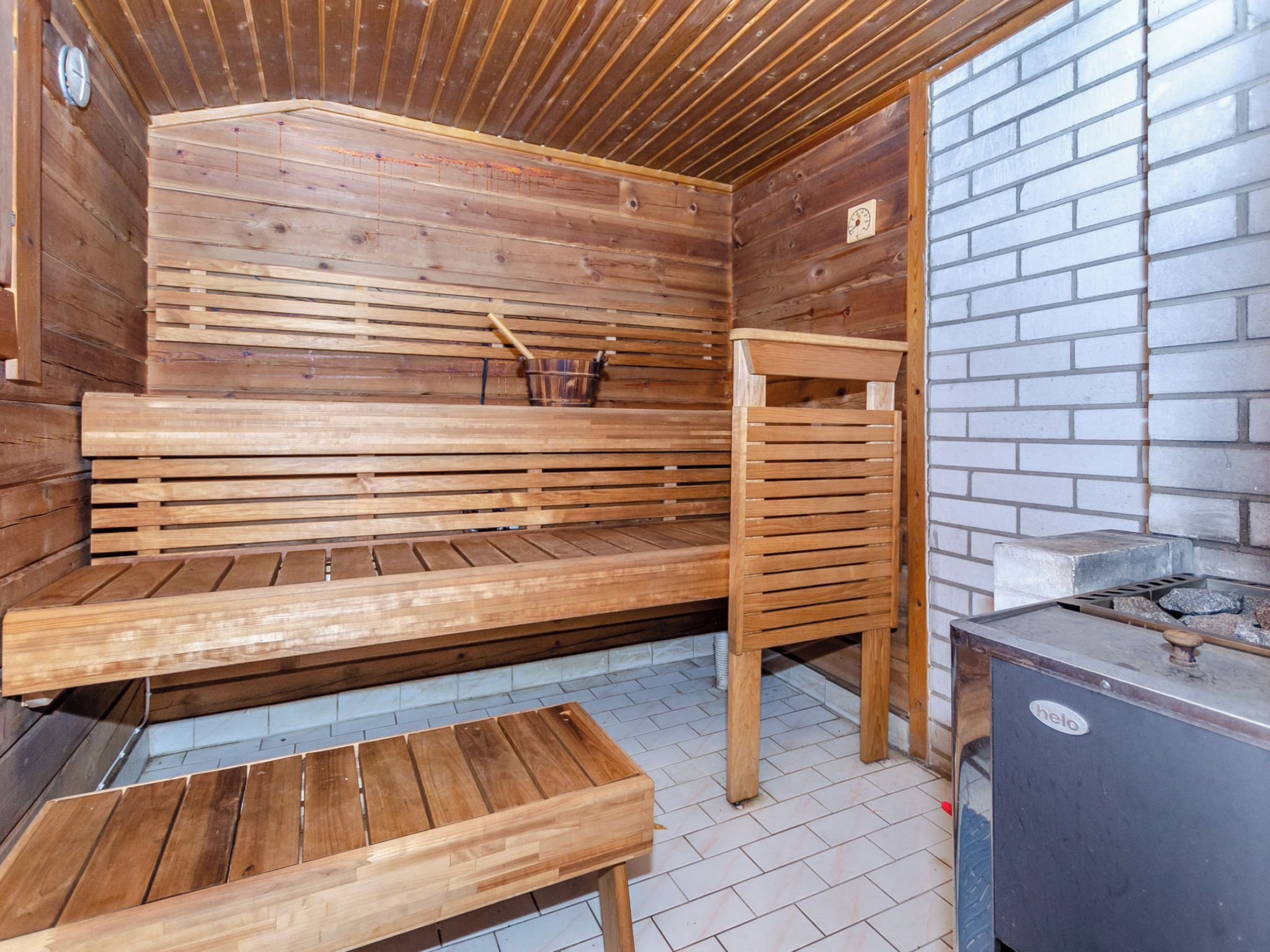 Una sala de sauna con bancos de madera, un banco con respaldo, un calentador de sauna y una pared de ladrillo.