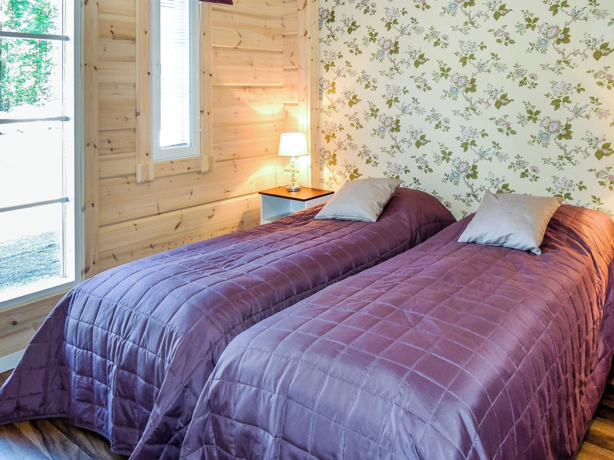 Un lit double avec une couvre-lit violet dans une chambre de style cabane, avec des murs en bois et un papier peint à motifs floraux.
