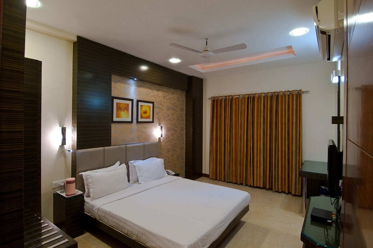 A hotel room with a bed, a nightstand, a desk with a computer, a ceiling fan, and a window with curtains.