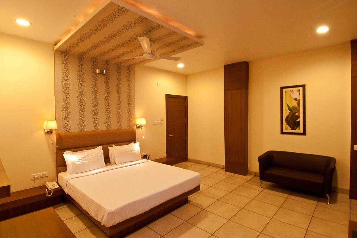 A hotel room with a large bed, a ceiling fan, a wooden headboard, a bedside lamp, a dark-colored sofa, and a framed picture on the wall.