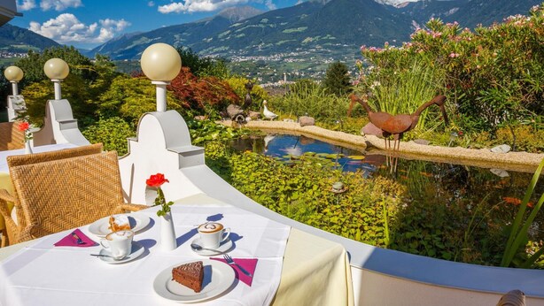 Una mesa dispuesta con un pastel, café y una rosa roja, con vistas a un jardín con un estanque y montañas al fondo.