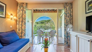 A room with a sofa, a dining table with chairs, a potted plant, a TV, and a balcony with a view of greenery. - Les Rivages de Coudoulière by Interhome (Six Fours La Coudoulière)