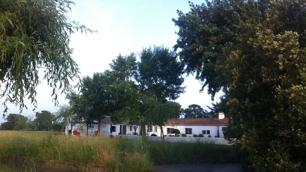 A white building with a red roof, surrounded by trees and a body of water.