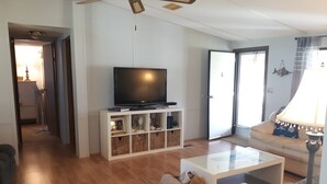 A living room with a flat-screen TV on a white stand, a glass coffee table, a sofa, and a lamp. - The Hoi Toider @ Harkers Island (Harkers Island)