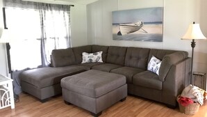 A modern living room with a sectional sofa, a matching ottoman, a lamp, and a painting of a boat on water. - The Hoi Toider @ Harkers Island (Harkers Island)
